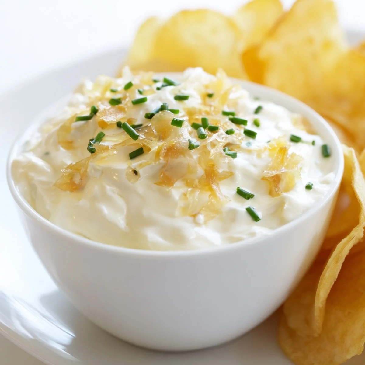 Bowl of creamy caramelized onion dip garnished with fresh chives alongside crispy golden homemade potato chips.